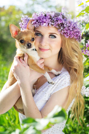 Beautiful blonde teen girl is wearing wreath of lilac flowers and small chuhuahua dogの写真素材