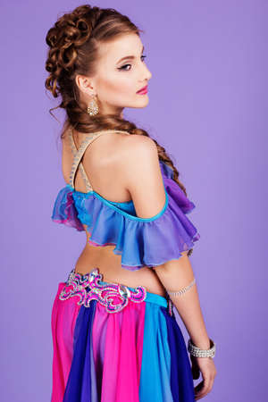 Beautiful belly dancer girl with nice makeup and heardress is wearing a colorful fashion costume. Isolated on purpleの写真素材