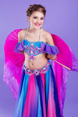 Beautiful belly dancer girl is wearing a colorful fashion costume and holding fan. Isolated on purpleの写真素材