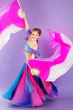 Beautiful belly dancer girl is wearing a colorful fashion costume and holding fan. Isolated on purpleの写真素材