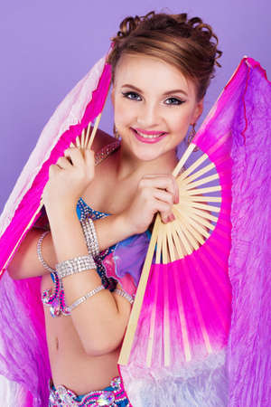 Beautiful belly dancer girl is wearing a colorful fashion costume and holding fan. Isolated on purpleの写真素材