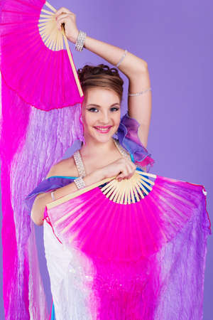 Beautiful belly dancer girl is wearing a colorful fashion costume and holding fan. Isolated on purpleの写真素材