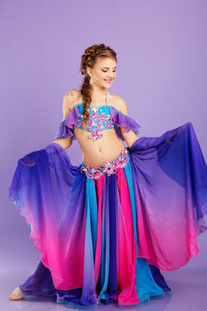 Beautiful belly dancer girl with nice makeup and heardress is wearing a colorful fashion costume. Isolated on purpleの写真素材