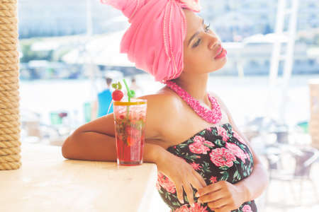 Young south african zulu woman is sitting near bar and drinking mojito, summer time with fashion makeup is wearing pink turban fashion dress and beadの写真素材