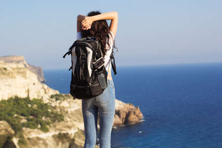 Back view of young pretty slim woman standing with backpack on cliff's edge and looking into oceanの写真素材