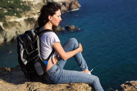 Pretty traveler girl is sitting on rock edge over sea background  with backpack, summer timeの写真素材