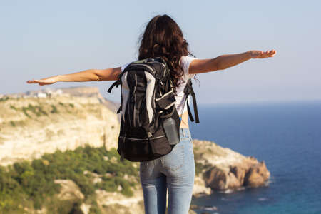 Back view of young pretty slim woman standing with backpack on cliff's edge and looking into a wide valleyの写真素材