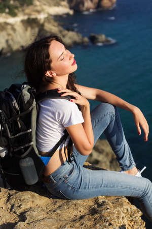 Pretty traveler girl is sitting on rock edge over sea view with backpack, summer timeの写真素材