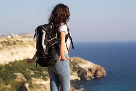 Back view of young pretty woman standing with backpack on cliff's edge and looking into a wide valleyの写真素材