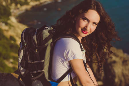 Pretty smiling traveler girl backpacker is drinking water on rock peak over sea view with backpack, summer vacationsの写真素材