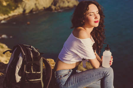 Pretty traveler girl backpacker is drinking water on rock peak over sea view with backpack, summer vacationsの写真素材