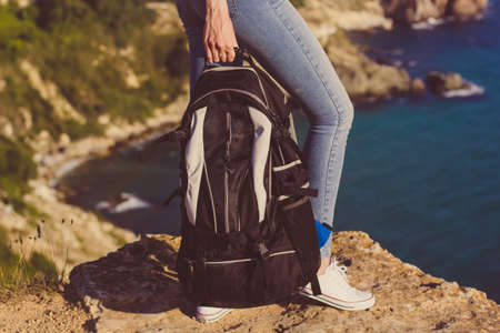 Pretty traveler girl backpacker is holding backpack on rock peak over sea view, summer vacationsの写真素材