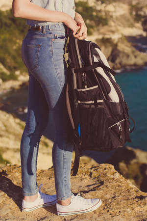 Pretty traveler girl backpacker is holding backpack on rock peak over sea view, summer vacationsの写真素材