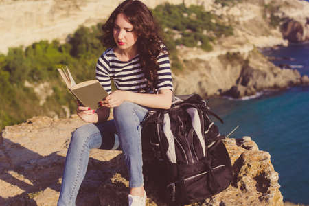 Pretty traveler brunette girl is sitting on rock and reading book over sea view with backpack, summer timeの写真素材