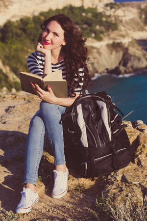 Pretty traveler brunette girl is sitting on mountain peak and reading book over sea view with backpack, summer timeの写真素材