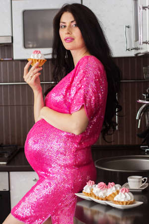 Pregnant happy woman is wearing fashion pink dress in kitchen eating sweet cakesの写真素材
