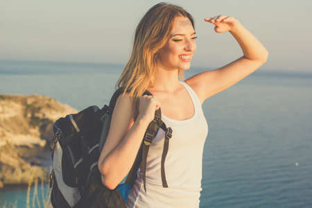 Pretty traveler girl is standing on rock edge over sea view with backpack, summer timeの写真素材