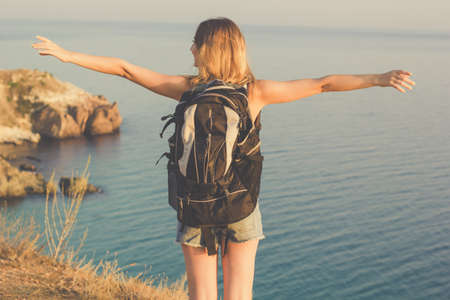 Back view traveler girl is standing on rock edge with backpack over sea view with backpack, summer timeの写真素材