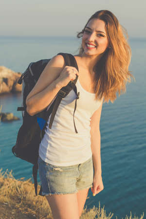 Pretty traveler girl is standing on rock edge over sea view with backpack, summer vacationsの写真素材