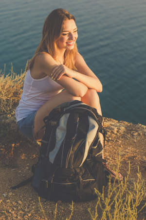 Pretty traveler girl is sitting on rock cliff over sea view with backpack, summer vacationsの写真素材