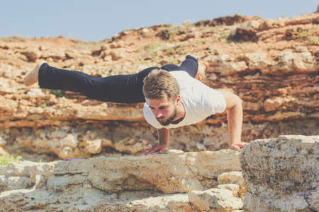 Young sportive man is practicing acroyoga exercises in the early morning on the beach with stonesの写真素材