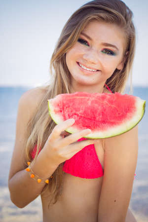 Beauty smiling teenager girl is wearing pink bikini and eating watermelon near seaの写真素材