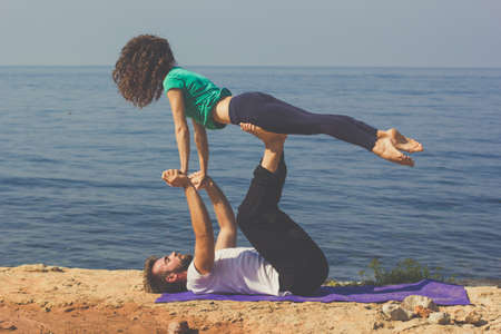 Young sportive couple male and female are practicing acroyoga exercises in the early morning on the beach over sea backgroundの写真素材