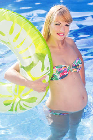 Beautiful pregnant woman is holding green rubber ring in blue water of swimming pool, summer vacationsの写真素材