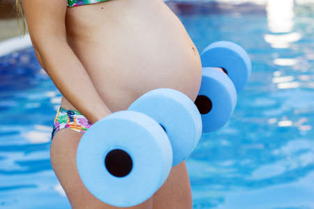 Beautiful pregnant woman is water aerobics with blue lightweight dumbbells near swimming poolの写真素材