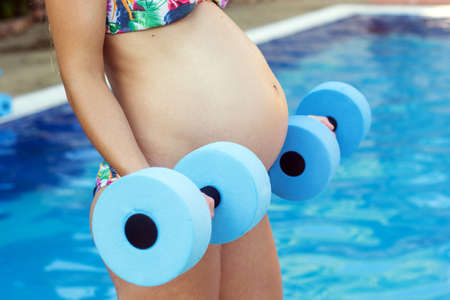 Beautiful pregnant woman is doing sport exercises with blue lightweight dumbbells near swimming poolの写真素材