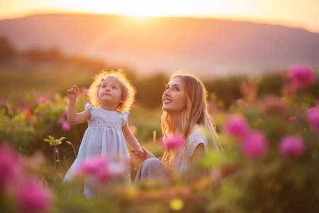 Beautiful child girl with young happy mother are wearing casual clothes walking in roses garden over sunset lightsの写真素材