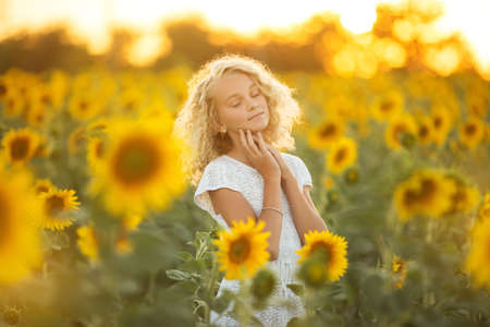 Pretty curly girl in white dress in filed of sunflowers over sunset lightsの写真素材