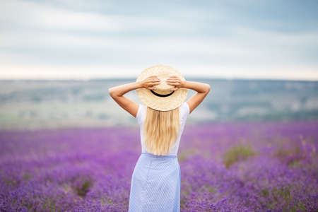 Beautiful blonde girl on the lavender field is holding hatの写真素材