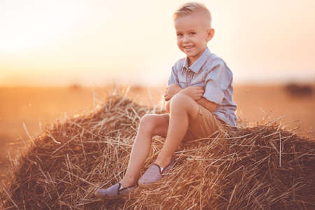 Pretty smiling child boy is wearing casual clothes near on stack of hay in the fieldの写真素材