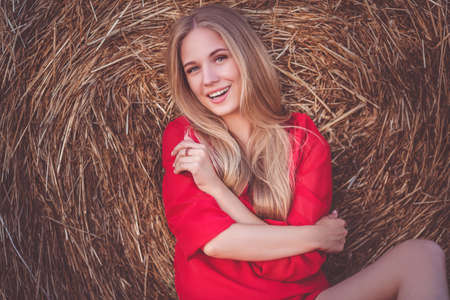 Pretty girl is sitting near stack of hay in the fieldの写真素材