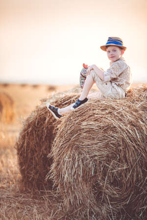 Pretty ginger child boy is wearing casual clothes near on stack of hay in the fieldの写真素材