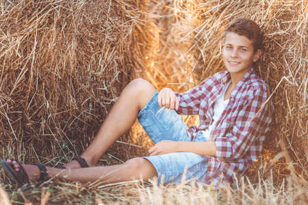 Pretty smiling teen boy is wearing casual clothes near on stack of hay in the fieldの写真素材