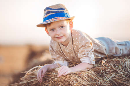 Pretty ginger child boy is wearing casual clothes and hat near on stack of hay in the fieldの写真素材