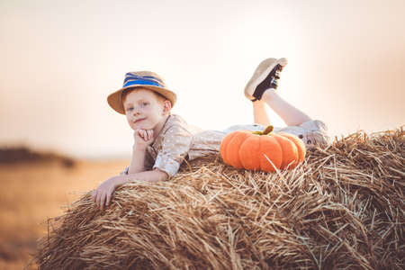 Pretty ginger child boy is wearing casual clothes and hat near on stack of hay in the fieldの写真素材