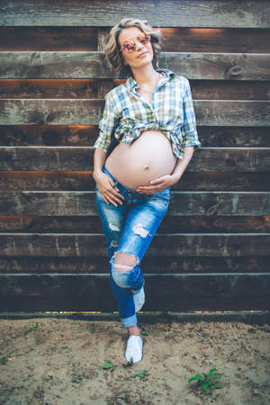 Beautiful smiling pregnant girl is wearing casual clothes and sunglasses is posing over wooden wallの写真素材