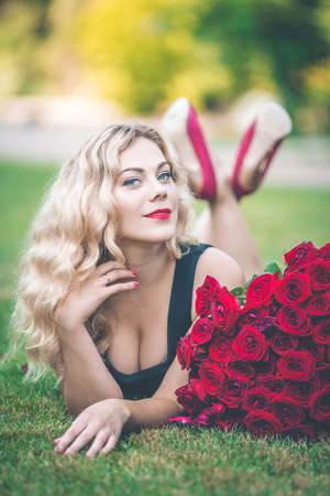 Elegant blonde woman lying on green lawn with big bouquet of 101 red rosesの写真素材
