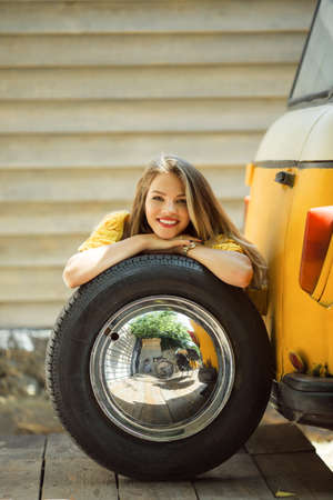 Happy smiling girl is wearing yellow sweater is posing with auto wheel near old retro bus, autumn conceptの写真素材