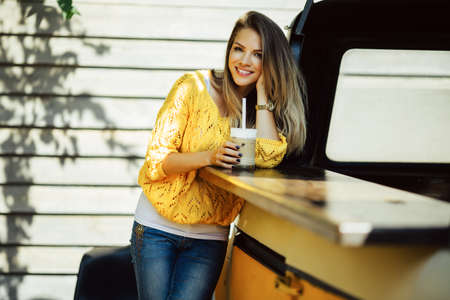 Pretty smiling woman is wearing yellow sweater drinking coffee latte near old retro bus, food and drink conceptの写真素材
