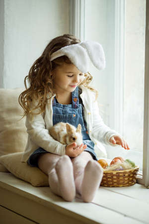 Cute happy little child girl is wearing bunny ears on Easter day sitting on window sill holding her friend little colorful rabbitの写真素材