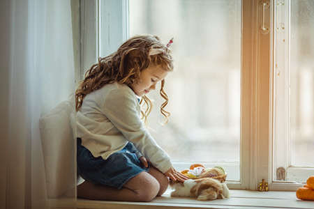 Beautiful happy child girl is sitting on window sill with her friend little colorful rabbit, Easter holiday conceptの写真素材
