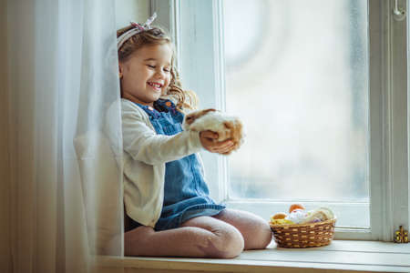 Beautiful happy curly child girl is sitting on window sill with her friend little colorful rabbit, Easter holiday conceptの写真素材
