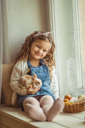 Pretty happy child girl is sitting on window sill with her friend little colorful rabbit, Easter holiday conceptの写真素材