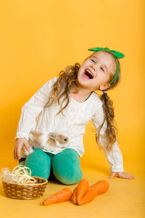 Pretty happy child girl with carrots and her friend little colorful rabbit, Easter holiday concept isolated on yellowの写真素材