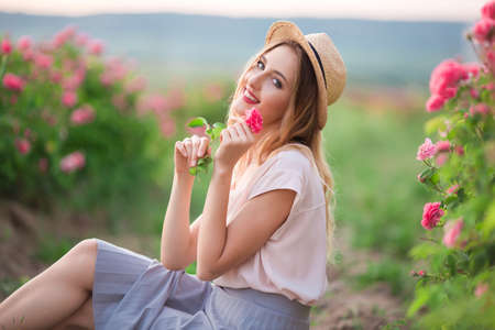 Beautiful young girl is wearing hat having rest in a garden with pink blossom rosesの写真素材