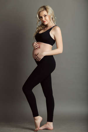 Pregnant beautiful blonde woman is wearing sportive black clothes isolated on gray, healthy life conceptの写真素材